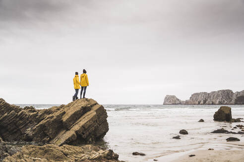 Young woman wearing yellow rain jackets and standing on rock at the beach, Bretagne, France - UUF19685