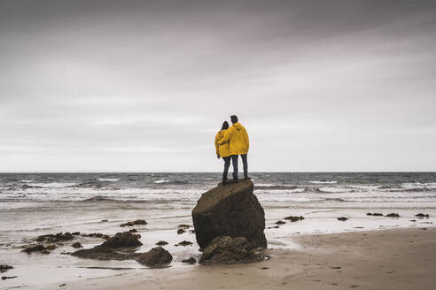 Junge Frau in gelben Regenjacken, die auf einem Felsen am Strand steht, Bretagne, Frankreich - UUF19686