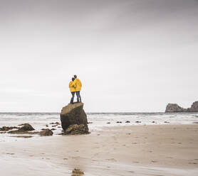 Junge Frau in gelben Regenjacken, die auf einem Felsen am Strand steht, Bretagne, Frankreich - UUF19687