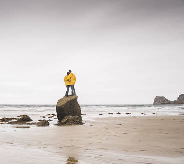 Young woman wearing yellow rain jackets and standing on rock at the beach, Bretagne, France - UUF19687