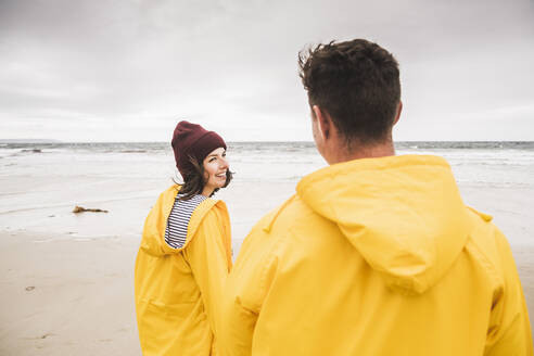 Young woman wearing yellow rain jackets and walking along the beach, Bretagne, France - UUF19689