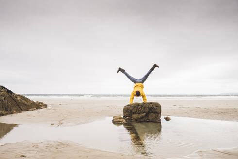 Junger Mann mit gelber Regenjacke am Strand, der einen Handstand auf einem Felsen macht, Bretagne, Frankreich - UUF19691