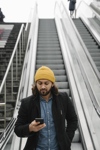 Porträt eines bärtigen Mannes, der auf einer Rolltreppe steht und auf sein Smartphone schaut, lizenzfreies Stockfoto