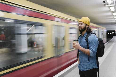 Porträt eines Mannes mit Rucksack und Kaffee zum Mitnehmen, der am Bahnsteig wartet, Berlin, Deutschland - AHSF01163