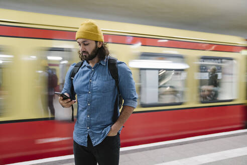 Bärtiger Mann mit Rucksack schaut auf sein Smartphone, während er am Bahnsteig wartet, Berlin, Deutschland - AHSF01166