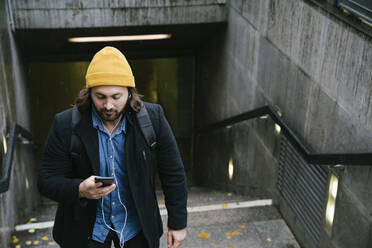 Man with earphones leaving train station on rainy day looking at cell phone - AHSF01173