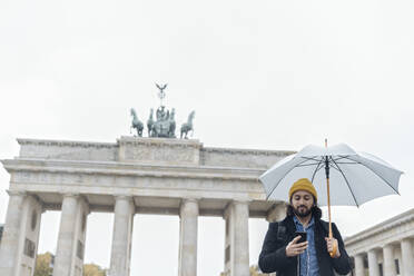 Mann mit Regenschirm und Smartphone vor dem Branderburger Tor, Berlin, Deutschland - AHSF01175