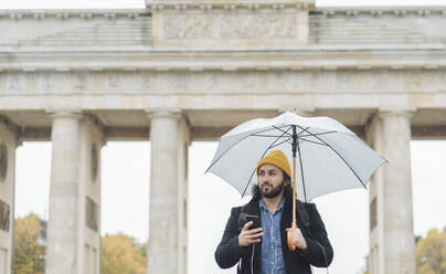 Mann mit Regenschirm und Smartphone vor dem Branderburger Tor, Berlin, Deutschland - AHSF01176