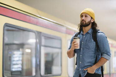 Porträt eines Mannes mit Rucksack und Kaffee zum Mitnehmen, der am Bahnsteig wartet, Berlin, Deutschland - AHSF01181