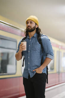 Porträt eines Mannes mit Rucksack und Kaffee zum Mitnehmen, der am Bahnsteig wartet, Berlin, Deutschland - AHSF01182