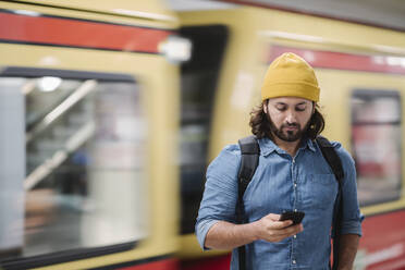 Porträt eines Mannes mit Rucksack, der auf sein Smartphone schaut, während er am Bahnsteig wartet, Berlin, Deutschland - AHSF01184