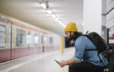 Mann mit Rucksack hört Musik mit Smartphone und Kopfhörern beim Warten am Bahnsteig, Berlin, Deutschland - AHSF01186
