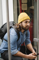 Man with backpack listening music with smartphone and earphones in commuter line, Berlin, Germany - AHSF01188