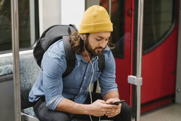 Man with backpack listening music with smartphone and earphones in commuter line, Berlin, Germany - AHSF01189