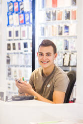 Porträt eines lächelnden jungen männlichen Auszubildenden, der ein Mobiltelefon repariert, während er an einem beleuchteten Schreibtisch in einem Geschäft sitzt - MASF14332