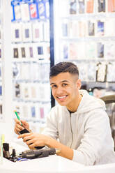 Porträt eines lächelnden jugendlichen Auszubildenden, der ein Mobiltelefon repariert, während er an einem beleuchteten Schreibtisch in einem Geschäft sitzt - MASF14334