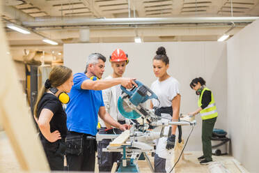 Auszubildende beobachten einen selbstbewussten männlichen Tischler beim Schneiden mit der Elektrosäge in der Werkstatt - MASF14365