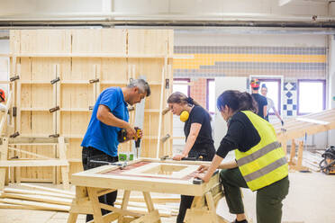 Young female trainees looking at male instructor using power drill on plank at workbench in workshop - MASF14366