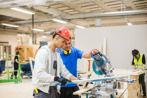 Teenage carpenter using level while standing by instructor with electric saw at illuminated workshop - MASF14367