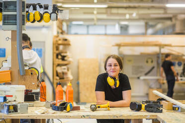 Porträt einer lächelnden jungen Schreinerin, die sich in einer beleuchteten Werkstatt auf eine Werkbank mit Elektrowerkzeugen stützt - MASF14378