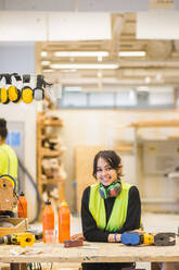 Porträt eines lächelnden jungen weiblichen Auszubildenden, der sich in einer Werkstatt an eine Werkbank lehnt - MASF14386