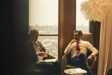 Männliche Finanzberater, die in einer Anwaltskanzlei am Fenster sitzend diskutieren - MASF14452