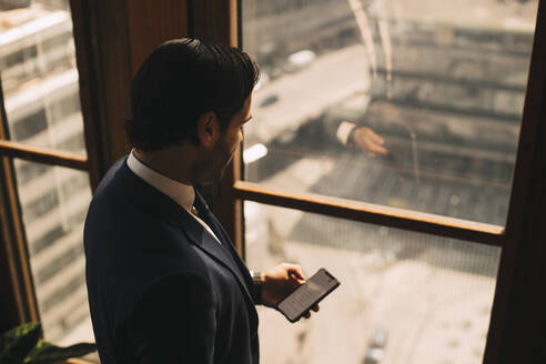 Hoher Blickwinkel eines männlichen Anwalts, der sein Smartphone am Fenster im Büro benutzt - MASF14459