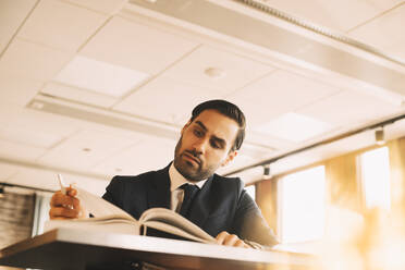 Niedriger Blickwinkel eines Finanzberaters, der ein Buch am Tisch im Büro liest - MASF14528