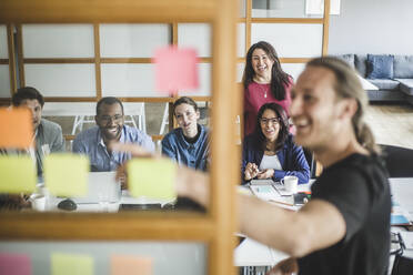 Lächelnde Kollegen schauen auf einen Geschäftsmann, der in einer Sitzung durch eine Glaswand hindurch die Strategie erklärt - MASF14554