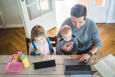 Hoher Blickwinkel des Vaters, der am Laptop arbeitet, während er mit seinem niedlichen Sohn bei der Tochter sitzt und ein digitales Tablet am Tisch benutzt - MASF14605
