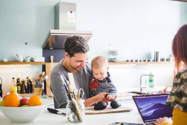 Smiling man carrying son holding tomato on cutting board at kitchen island - MASF14610