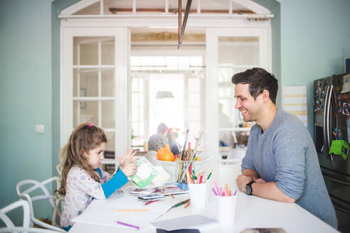 Lächelnder Vater betrachtet ein Mädchen, das ein Bilderbuch in der Küche hält - MASF14618