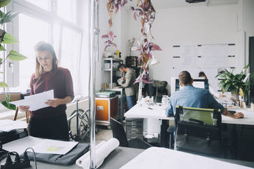 Kreative Geschäftsfrau, die Papiere prüft, während Kollegen im Hintergrund im Büro arbeiten - MASF14630