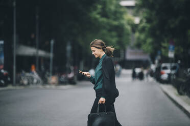 Confident businesswoman using smart phone while crossing street in city - MASF14642
