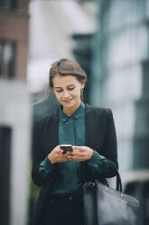 Confident businesswoman text messaging through smart phone while walking in city - MASF14646