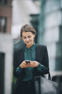Selbstbewusste Geschäftsfrau, die beim Spazierengehen in der Stadt eine Textnachricht über ihr Smartphone verschickt - MASF14646
