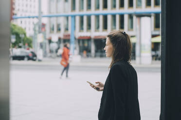 Seitenansicht einer zuversichtlichen Geschäftsfrau, die ein Mobiltelefon hält, während sie auf der Straße in der Stadt spazieren geht - MASF14651