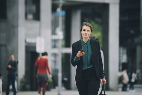 Porträt einer selbstbewussten Geschäftsfrau, die ihr Smartphone in der Hand hält, während sie auf einer Straße in der Stadt spazieren geht, lizenzfreies Stockfoto