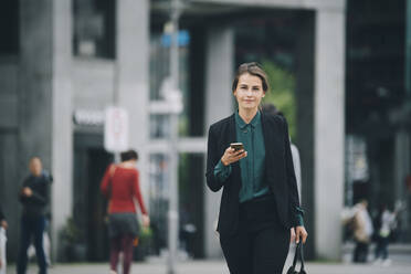 Portrait of confident businesswoman holding smart phone while walking on street in city - MASF14653