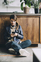 Full length of creative businesswoman using mobile phone while sitting against kitchen island in office - MASF14657