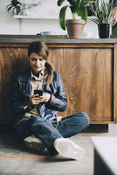 Kreative Geschäftsfrau in voller Länge, die ein Mobiltelefon benutzt, während sie an der Kücheninsel im Büro sitzt - MASF14657