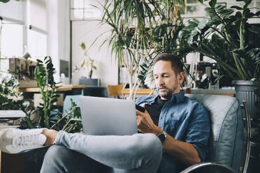 Selbstbewusster kreativer Geschäftsmann, der ein Mobiltelefon benutzt, während er mit seinem Laptop auf dem Sitz im Büro sitzt - MASF14661