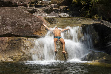 Young adult man sitting over rock and bathing on rainforest waterfall - CAVF69070