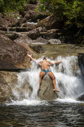 Young adult man sitting over rock and bathing on rainforest waterfall - CAVF69071