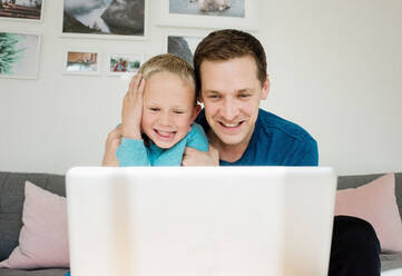 Vater und Sohn lachen zu Hause, während sie auf einen Laptop schauen - CAVF69170