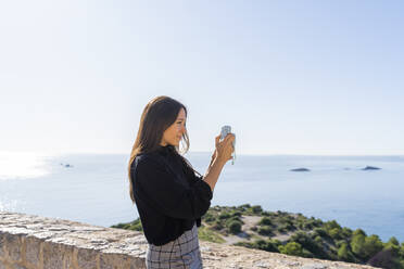 Porträt einer jungen brünetten Frau, die mit einer blauen Kamera von einem Aussichtspunkt in Ibiza aus fotografiert - AFVF04192