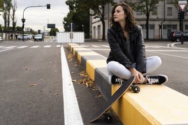 Junge Frau mit Skateboard, die auf einem Poller sitzt und in die Ferne schaut - GIOF07682
