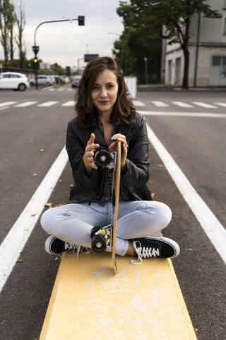 Porträt einer lächelnden jungen Frau mit Skateboard auf einem Poller sitzend, lizenzfreies Stockfoto