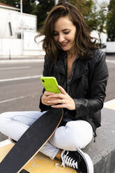 Lächelnde junge Frau mit Skateboard, die auf einem Poller sitzt und ein Smartphone benutzt - GIOF07685