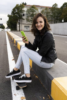 Porträt einer lächelnden jungen Frau mit Skateboard, die auf einem Poller sitzt und ein Smartphone benutzt - GIOF07687